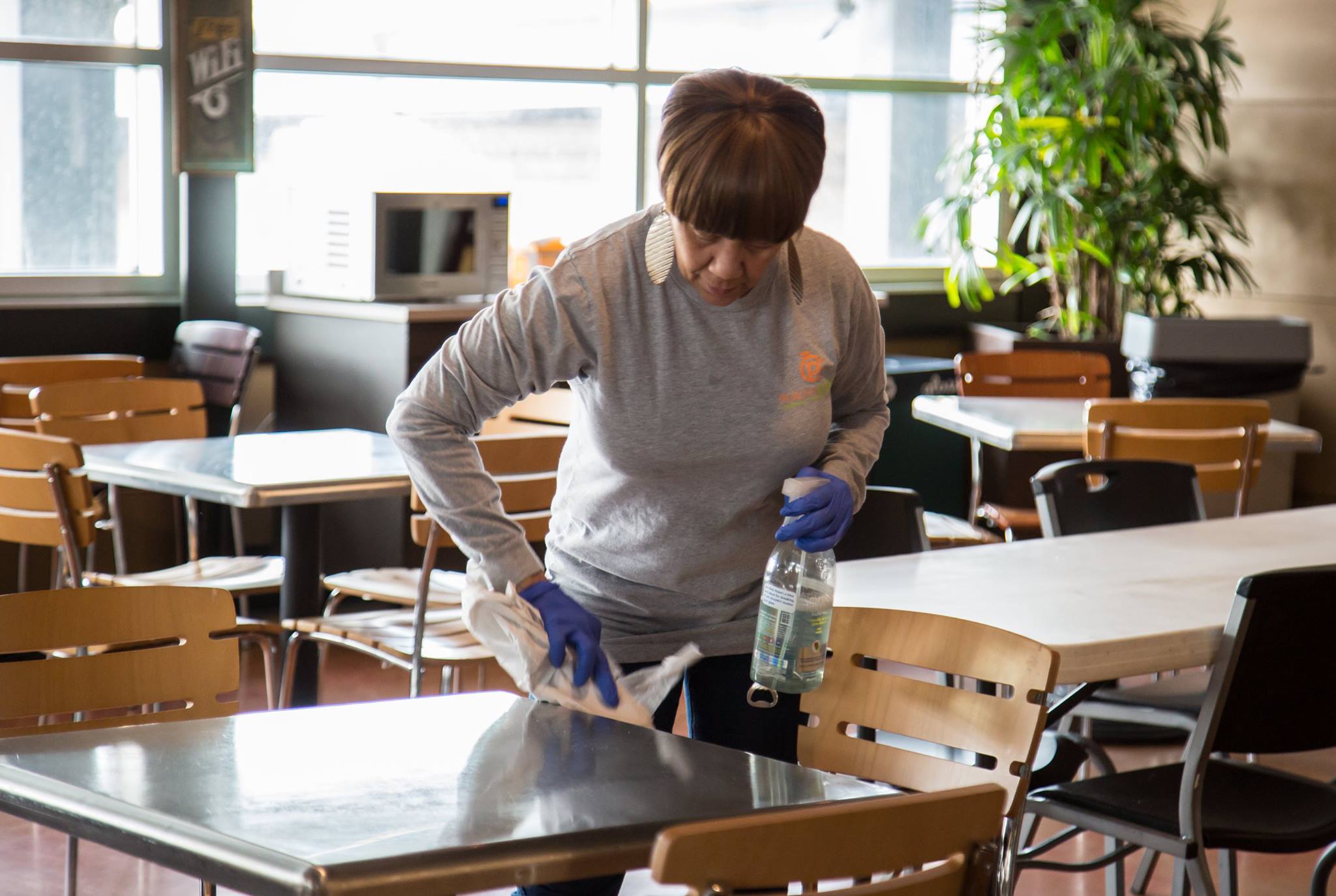 Professional cleaning services in action—worker wearing gloves sanitizes cafeteria table with spray and cloth in a shared dining area.